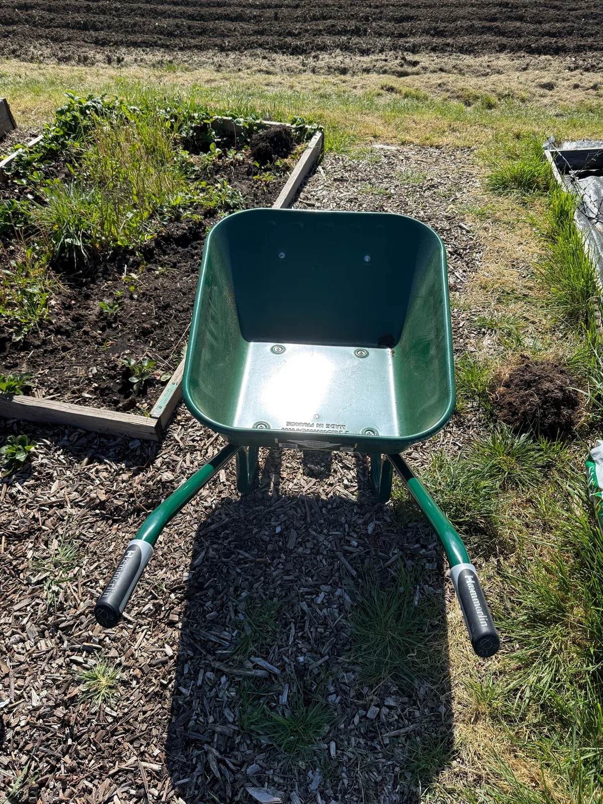 The empty green Haemmerlin wheelbarrow head-on, its name on the handle grips, on a woodchip path between the beds